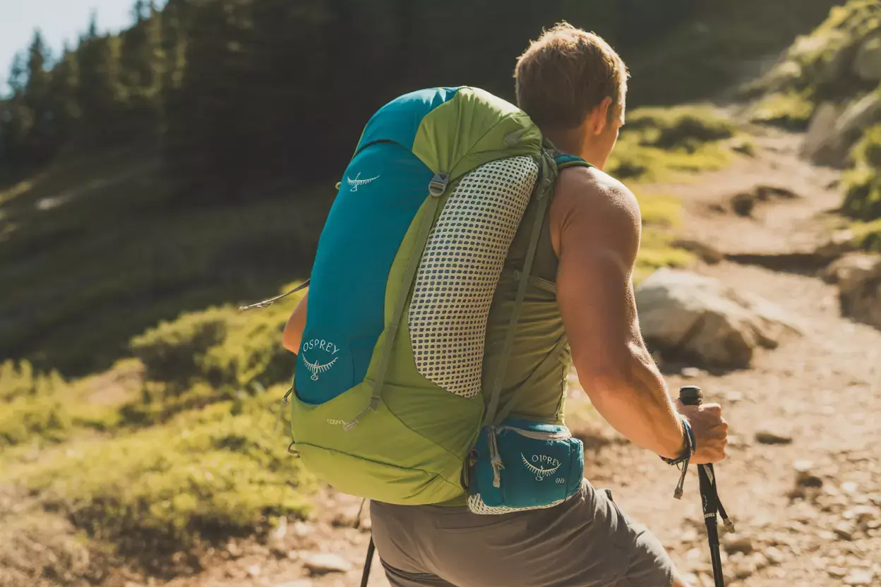 A hiker with a large green and blue backpack walks uphill on a sunny, rocky trail using trekking poles.