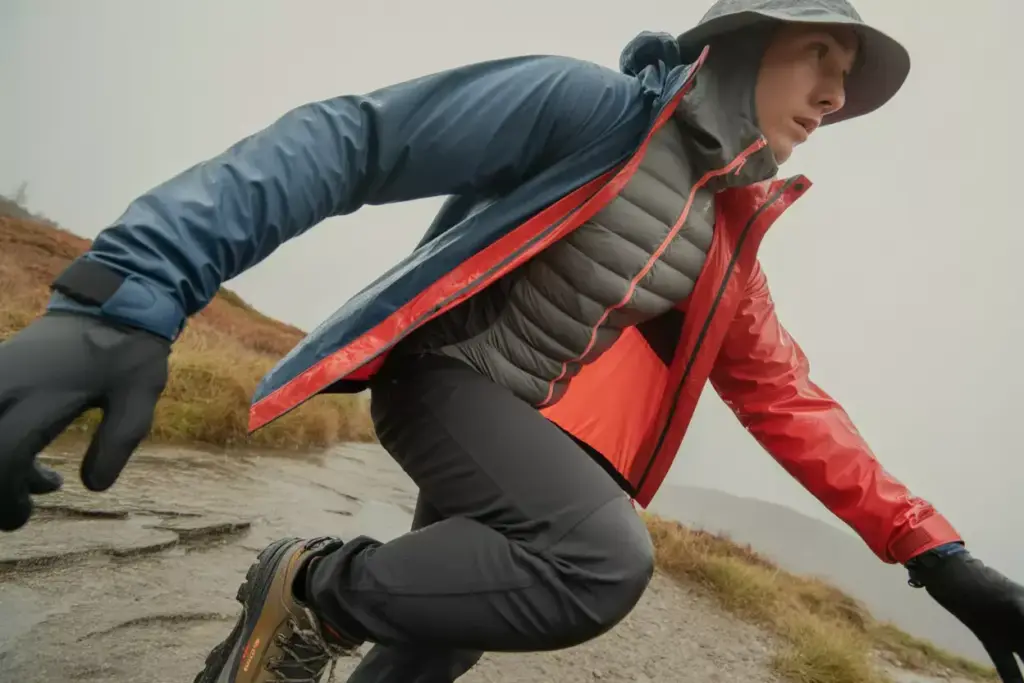 Person in outdoor gear crouching on a rocky trail, wearing gloves, boots, and a hooded jacket in rainy weather.