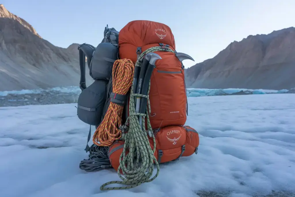 Orange hiking backpack with climbing gear, ropes, and ice axes sits on snow with mountains in the background.