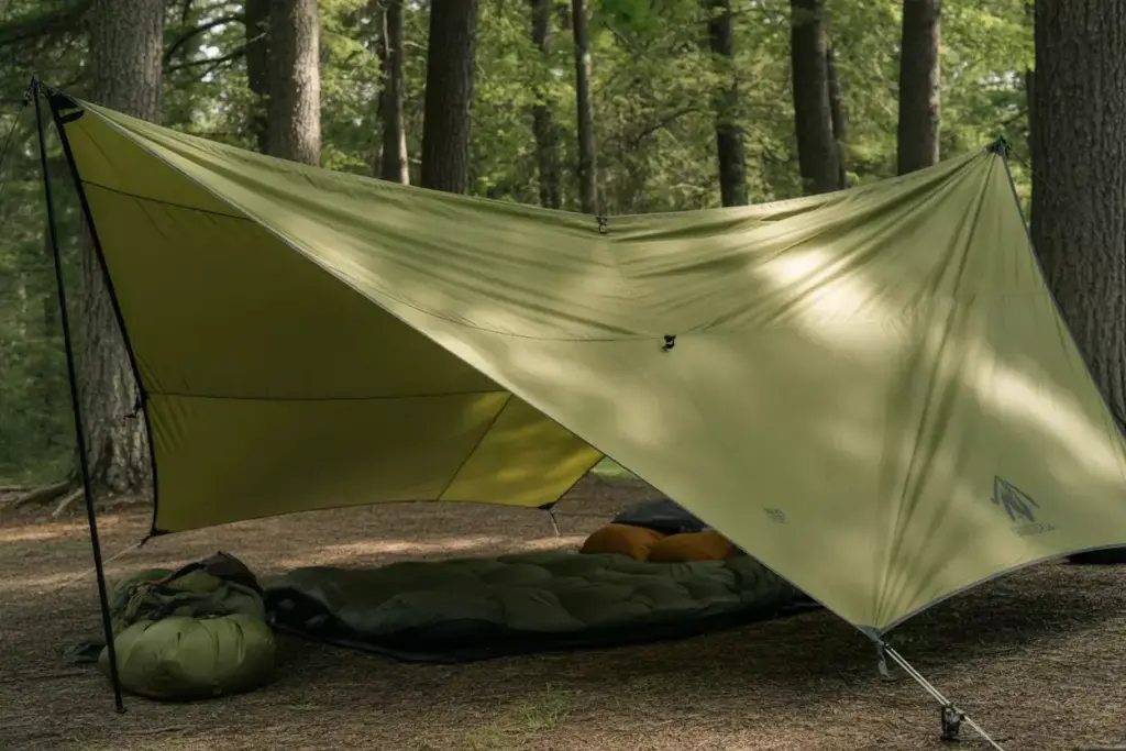 A green tarp shelter set up in a forest with sleeping bags and camping gear underneath.