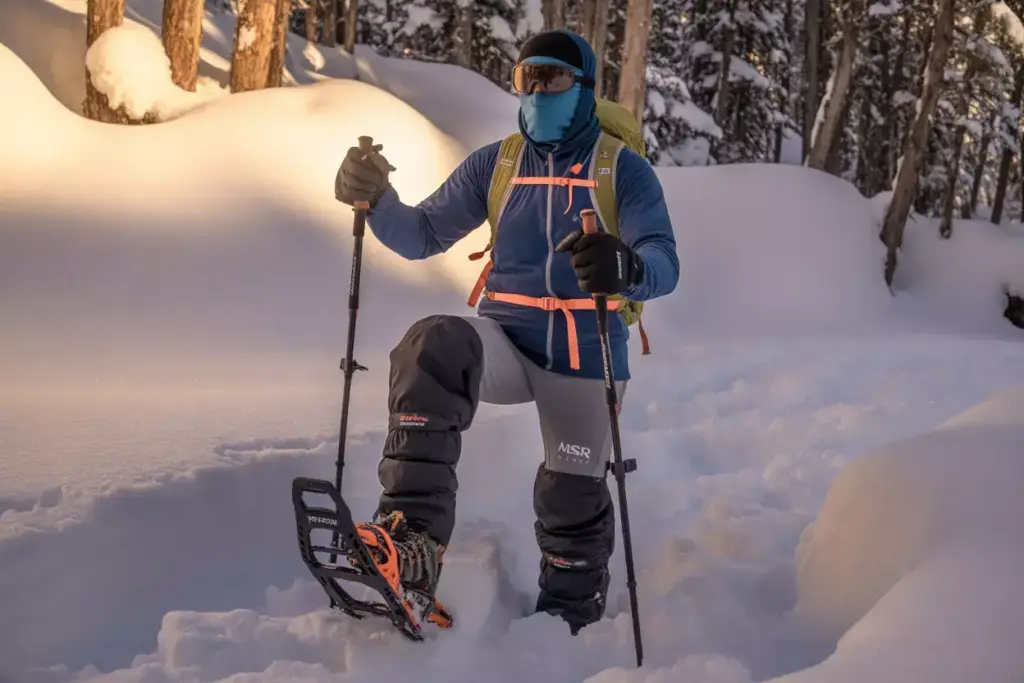 A person in winter gear snowshoeing through deep snow in a sunlit forest.
