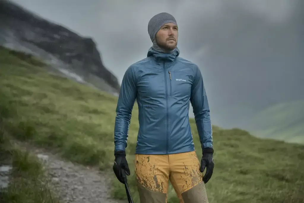 A man in outdoor gear stands on a grassy mountain trail, wearing a blue jacket and yellow shorts.