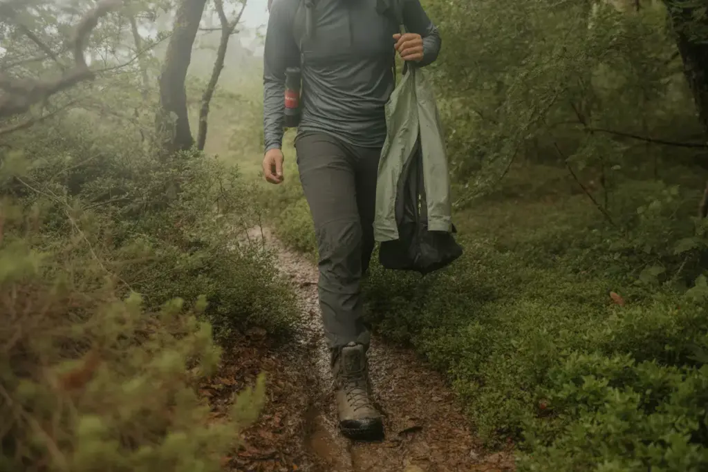 Person hiking on a muddy forest trail, wearing boots and carrying a jacket in one hand.
