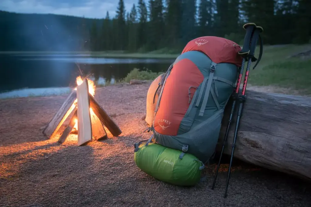 A camping backpack and hiking poles rest by a campfire beside a lake and forest at dusk.