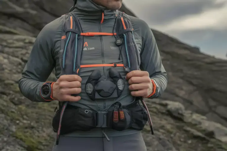 A person wearing a trail running vest and gear stands on a rocky landscape under a cloudy sky.