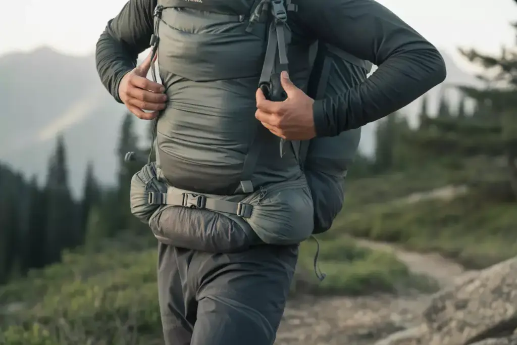 Close-up of a hiker adjusting a large gray backpack on a mountain trail with trees in the background.