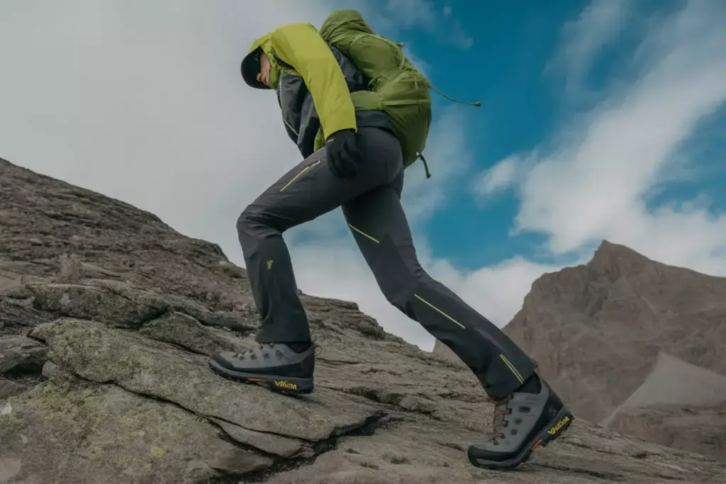 A hiker in outdoor gear climbs rocky terrain under a partly cloudy sky with mountains in the background.
