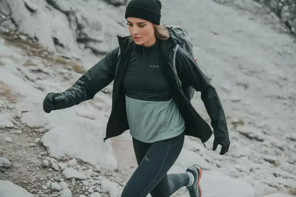 Woman hiking on rocky terrain wearing a black jacket, leggings, gloves, and a beanie.