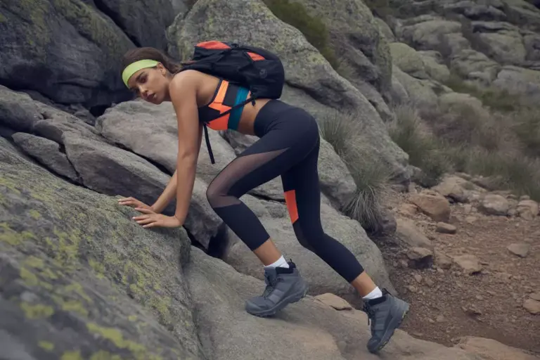 Woman in athletic wear climbs rocky terrain outdoors, wearing a backpack and headband.