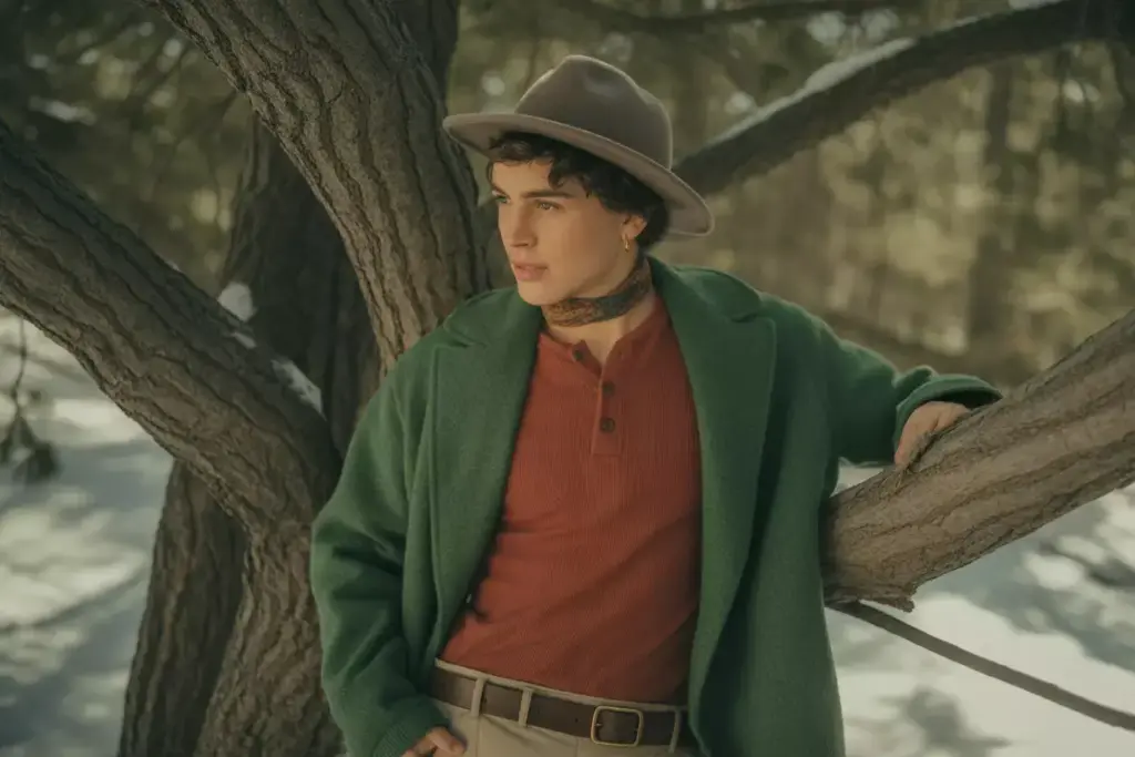 Person in a green coat and hat leans on a tree branch in a snowy outdoor setting.