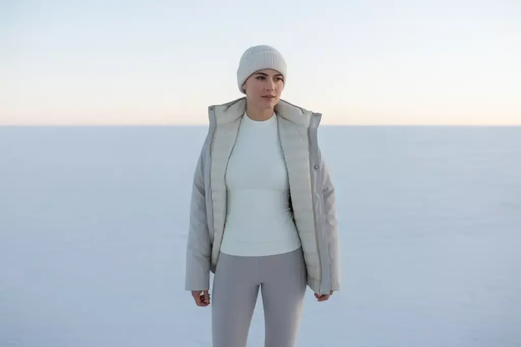 Woman in a white jacket, beanie, and leggings stands on a snowy plain under a pale sky.