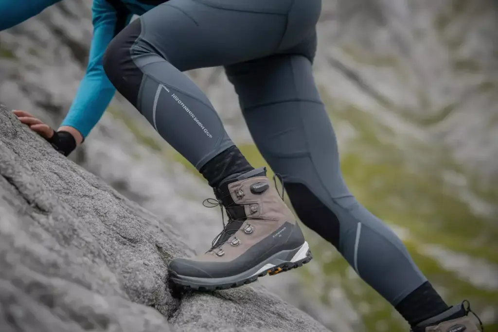 A person in hiking gear climbs a steep rocky surface, focusing on their boots and leggings.