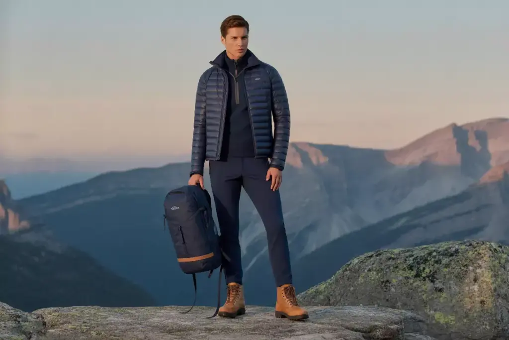 A man in outdoor gear stands on a rocky mountain with a backpack, overlooking distant cliffs at sunset.
