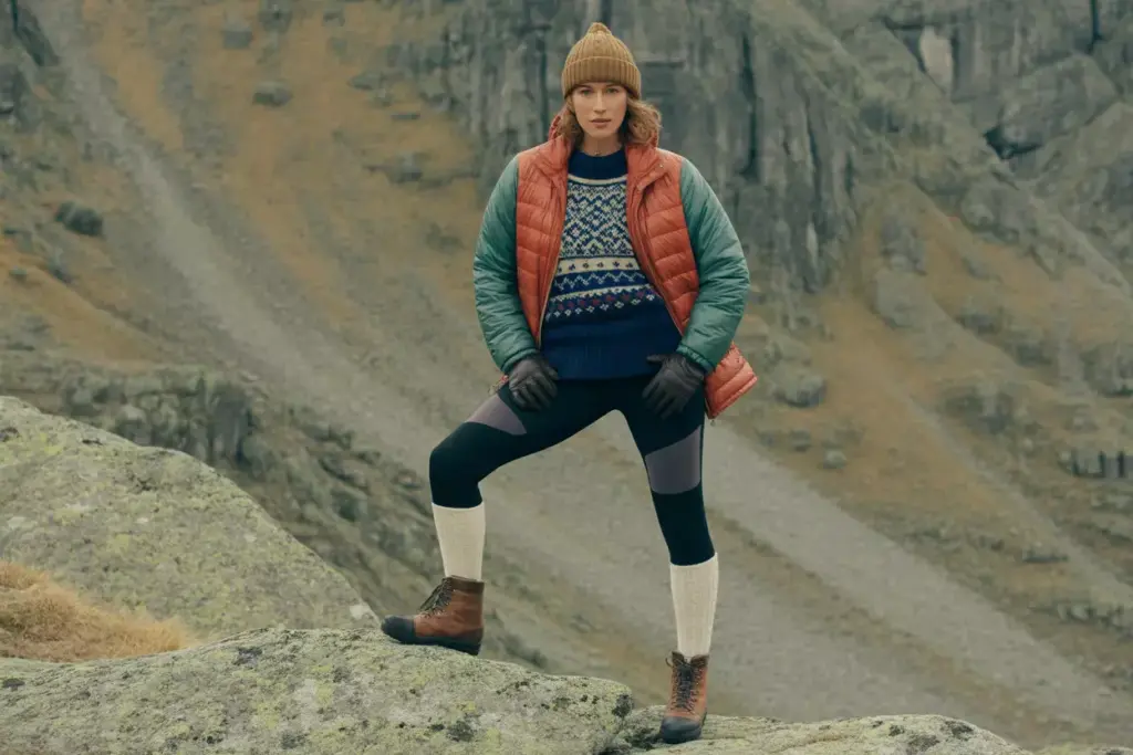 Person in winter outdoor clothing stands on a rocky slope with a mountain background.