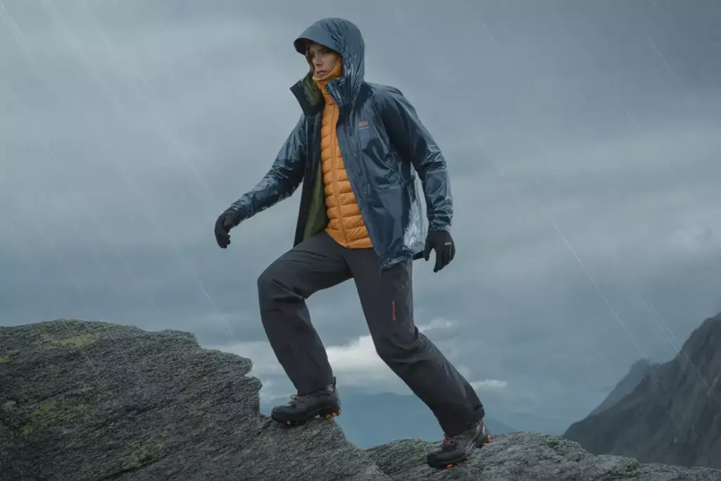 Person in outdoor gear climbing wet rocks in the rain, with a cloudy sky and mountains in the background.