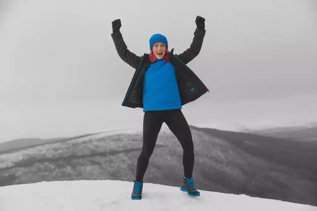 Person in blue winter clothes stands on snowy hill with arms raised, smiling against a foggy mountain background.