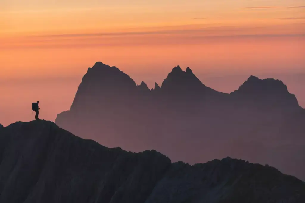 Mountain Range Silhouetted Against Vibrant Golden Hour Sky With Silhouetted Hiker On Ridge